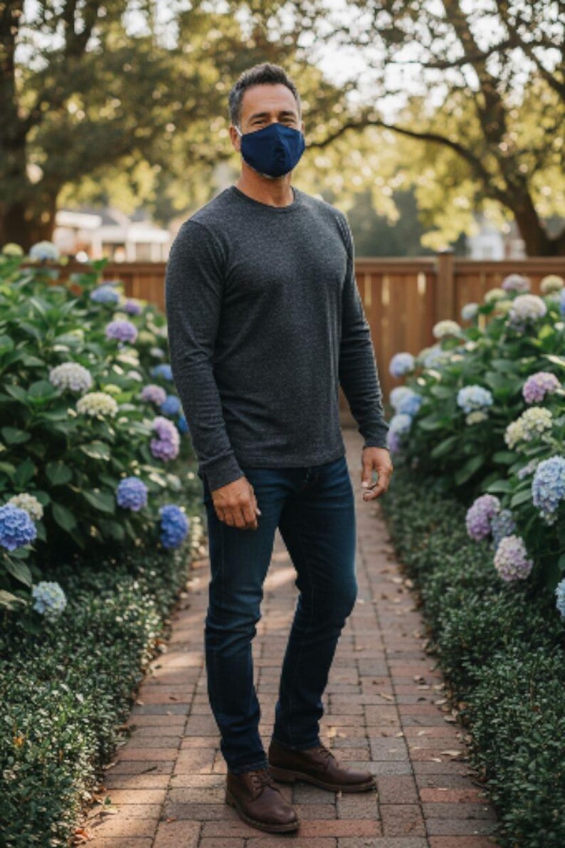 May include: A person wearing a navy blue face mask, dark grey long-sleeved top, and dark blue jeans. They are standing on a brick path lined with hydrangeas. Brown leather shoes complete the outfit.