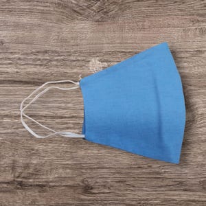 May include: A light blue face mask with white elastic ear loops. The mask is made of a solid, soft fabric and is designed to cover the nose and mouth. The mask is shown on a wooden surface.