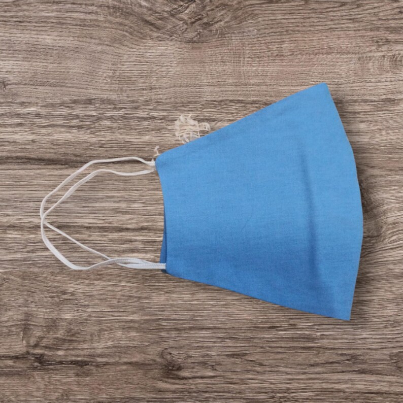 May include: A light blue face mask with white elastic ear loops. The mask is made of a solid, soft fabric and is designed to cover the nose and mouth. The mask is shown on a wooden surface.