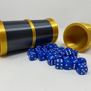 以下が含まれることがあります： 黒と金の円筒形のサイコロ入れ。白い点のある青いサイコロのコレクションが容器の近くに散らばっています。容器の長さは約10cmです。
