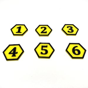 May include: Six yellow, hexagon-shaped game pieces with black borders and black numbers 1 through 6. The pieces are arranged on a white surface. The numbers are in a bold, sans-serif font.