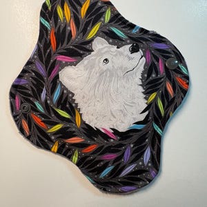 May include: A reusable menstrual pad with a white wolf illustration in the center, surrounded by a wreath of colorful leaves. The pad has a dark gray background with small white stars and a snap closure.