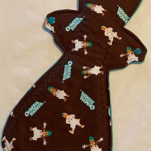 May include: Brown reusable menstrual pad with a teal trim and snap closure. The pad is patterned with white figures and the text "Oompa Loompa". The pad is designed for feminine hygiene.