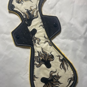 May include: Reusable cloth menstrual pad with a cream-colored octopus print on a black and yellow background. The pad has a contoured shape with snap closures for secure wear. The octopus design features black and brown tentacles.