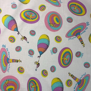 May include: White fabric featuring a repeating pattern of colorful Dr. Seuss illustrations. The design includes hot air balloons, swirling shapes, and characters, with text that reads "Oh, the Places You'll Go!" and "Dr. Seuss".