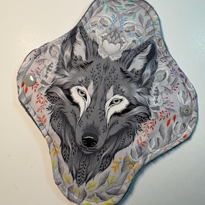 May include: A reusable cloth menstrual pad featuring a detailed gray wolf illustration. The pad has a gray floral pattern background with accents of orange, yellow, and blue. It has snap closures for secure wear.