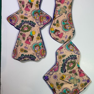 May include: Three reusable cloth pads with a pink background and a colorful print of cartoon children with the text "Cabbage Patch Kids" in a purple and white font. The pads have a purple trim.
