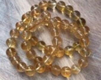 May include: A collection of golden-yellow, translucent citrine gemstone beads. The beads are round and strung together, forming multiple strands. The arrangement is on a wooden surface with a decorative border.