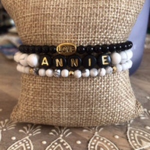 May include: A set of three bracelets with black and white beads. The top bracelet has black beads and a gold charm that says "LOVE". The middle bracelet has white beads and gold letter beads that spell "ANNIE". The bottom bracelet has white beads.