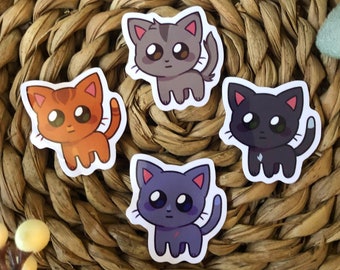 May include: Four colourful cat stickers. The stickers feature cartoon cats in orange, grey, black, and purple. Each cat has large eyes and a small tail. The stickers are on a woven, brown surface.