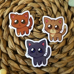 May include: Three cartoon cat stickers with different colored fur. The first cat is orange, the second is brown, and the third is black. All three cats have large eyes and are looking at the viewer.