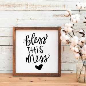 May include: A white wooden sign with a black handwritten message that says "bless this mess" with a small black heart below the text.