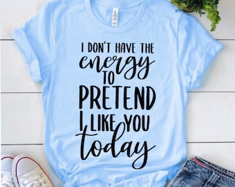 May include: A pale blue t-shirt featuring the black text: "I don't have the energy to pretend I like you today". The shirt is folded, with white trainers and denim shorts visible.