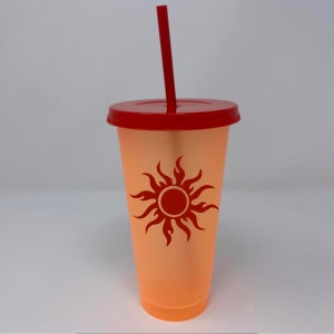 May include: A clear orange tumbler with a red lid and a red straw. The tumbler has a red sun with flames design on the front.