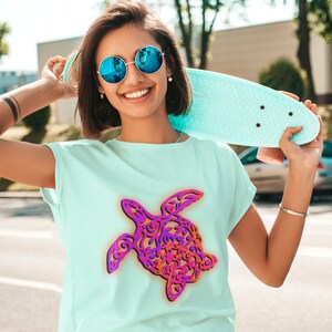 K&ouml;nnte beinhalten: Eine Frau tr&auml;gt eine blaue Sonnenbrille und ein hellblaues T-Shirt mit einem farbenfrohen, kunstvollen Meeresschildkr&ouml;ten-Design. Sie h&auml;lt ein hellblaues Skateboard.