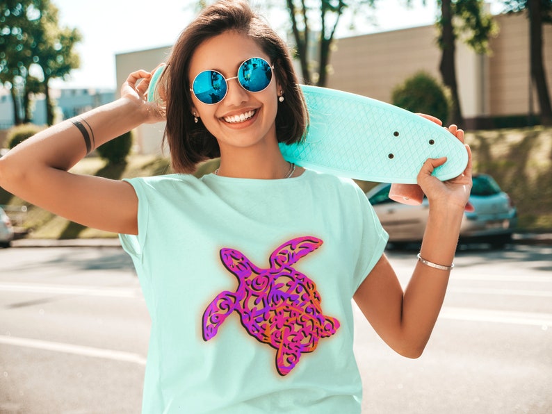 K&ouml;nnte beinhalten: Eine Frau tr&auml;gt eine blaue Sonnenbrille und ein hellblaues T-Shirt mit einem farbenfrohen, kunstvollen Meeresschildkr&ouml;ten-Design. Sie h&auml;lt ein hellblaues Skateboard.