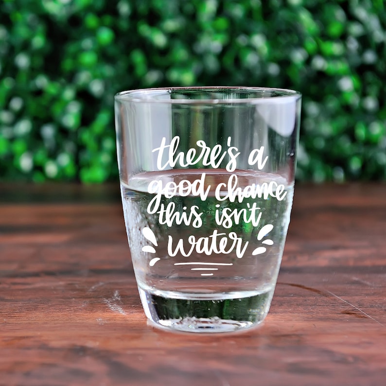 May include: A clear glass with a white inscription that reads "there's a good chance this isn't water". The glass is filled with a clear liquid and is sitting on a wooden surface.