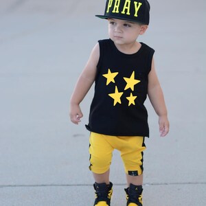 May include: A young boy wearing a black sleeveless shirt with yellow stars, yellow shorts with black stripes, black and yellow sneakers, and a black baseball cap with the word "PRAY" in yellow letters.