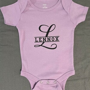May include: A purple baby onesie with a black letter L and the name Lennox printed on it.