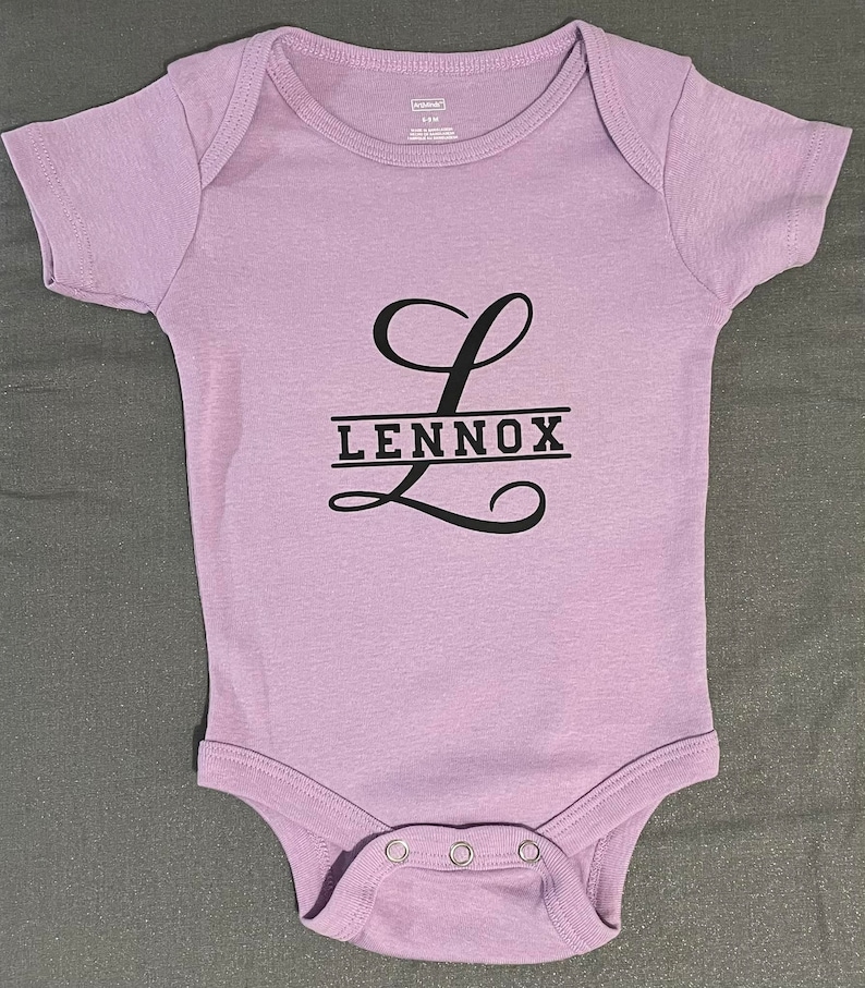 May include: A purple baby onesie with a black letter L and the name Lennox printed on it.