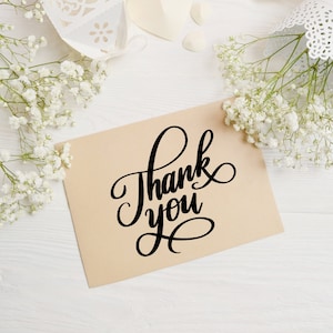 May include: A brown card with the text "Thank you" written in black cursive script. The card is surrounded by white baby's breath flowers.