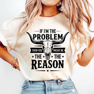 Peut inclure: Sweat-shirt crème avec un motif noir représentant un crâne de taureau et le texte "IF I'M THE PROBLEM THEN YOU MIGHT BE THE REASON". Le motif comprend des étoiles et un trait horizontal.
