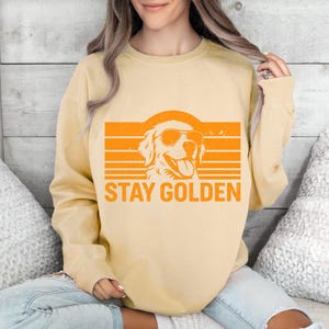May include: A light yellow sweatshirt with an orange graphic of a dog wearing sunglasses, set against an orange striped background. The text "STAY GOLDEN" is below the dog illustration. The sweatshirt is a casual style.