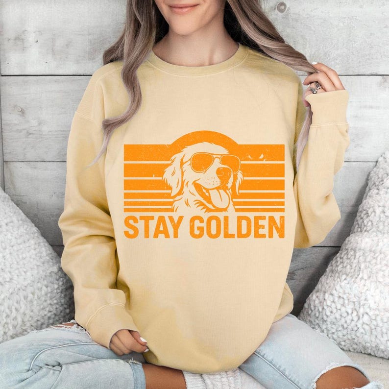 May include: A light yellow sweatshirt with an orange graphic of a dog wearing sunglasses, set against an orange striped background. The text "STAY GOLDEN" is below the dog illustration. The sweatshirt is a casual style.