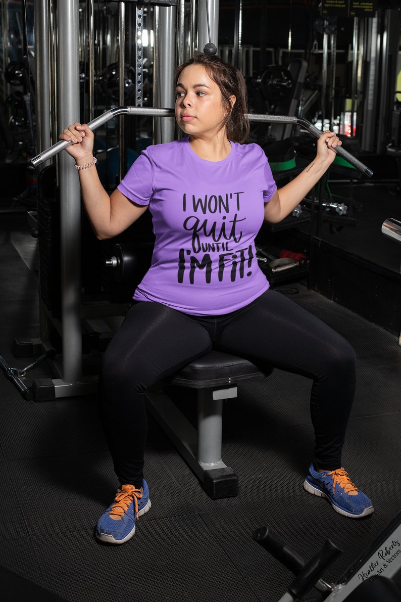 K&ouml;nnte beinhalten: Eine Frau im Fitnessstudio tr&auml;gt ein lila T-Shirt mit dem Text "I WON'T quit UNTIL I'M FIT!". Sie tr&auml;gt schwarze Leggings und blau-orangefarbene Turnschuhe. Sie benutzt ein Trainingsger&auml;t.