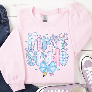 May include: A light pink sweatshirt with the words "First Grade" in a colorful, patterned design. The letters are decorated with floral, gingham, and bow accents. A blue bow and pencil graphic are at the bottom. A pair of purple sneakers and blue jeans are in the background.