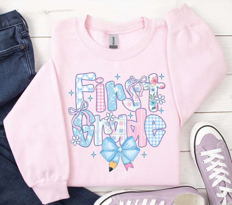 May include: A light pink sweatshirt with the words "First Grade" in a colorful, patterned design. The letters are decorated with floral, gingham, and bow accents. A blue bow and pencil graphic are at the bottom. A pair of purple sneakers and blue jeans are in the background.