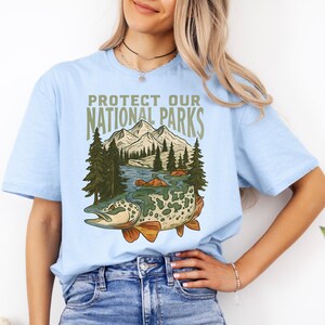 May include: Light blue t-shirt with the text "PROTECT OUR NATIONAL PARKS". The graphic features a fish, mountains, trees, and a river. The shirt is made of a soft material.