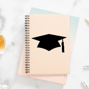 May include: A beige spiral notebook with a black graduation cap silhouette on the cover. The notebook is closed and sitting on a pink surface.