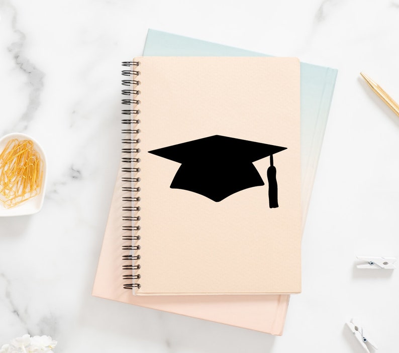May include: A beige spiral notebook with a black graduation cap silhouette on the cover. The notebook is closed and sitting on a pink surface.