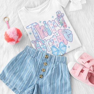 Puede incluir: Una camiseta blanca con las palabras "Third Grade" en un dise&ntilde;o colorido y estampado, combinada con pantalones cortos a rayas azules y blancas, zapatos rosas con lunares y un llavero con pomp&oacute;n rosa. Tambi&eacute;n se incluye un lazo blanco.