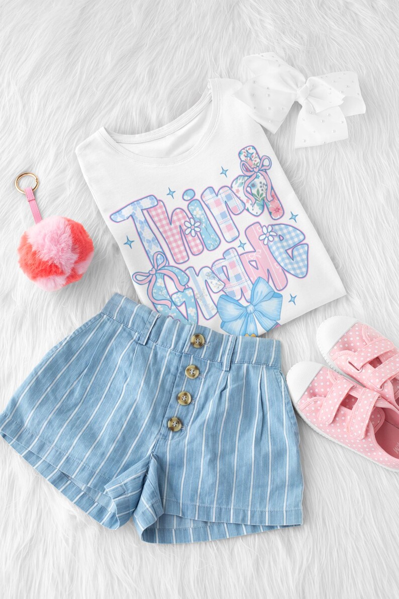 Puede incluir: Una camiseta blanca con las palabras "Third Grade" en un dise&ntilde;o colorido y estampado, combinada con pantalones cortos a rayas azules y blancas, zapatos rosas con lunares y un llavero con pomp&oacute;n rosa. Tambi&eacute;n se incluye un lazo blanco.