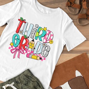 Puede incluir: Camiseta blanca con un gr&aacute;fico colorido que dice "Tercer Grado" con un arco&iacute;ris, l&aacute;pices y un globo terr&aacute;queo.