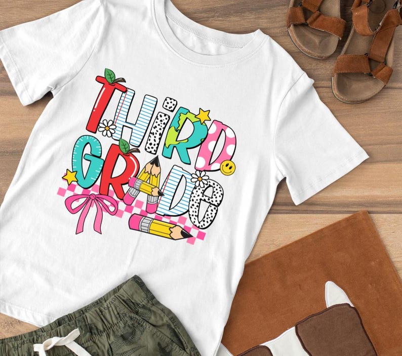 Puede incluir: Camiseta blanca con un gr&aacute;fico colorido que dice "Tercer Grado" con un arco&iacute;ris, l&aacute;pices y un globo terr&aacute;queo.