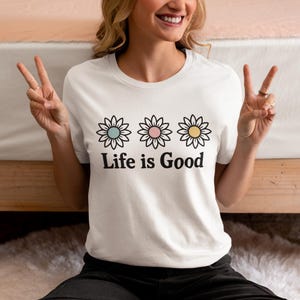 May include: White t-shirt with three daisy flower designs in pastel shades above the text "Life is Good". The flowers have blue, pink, and yellow centres. A casual, everyday wear item.