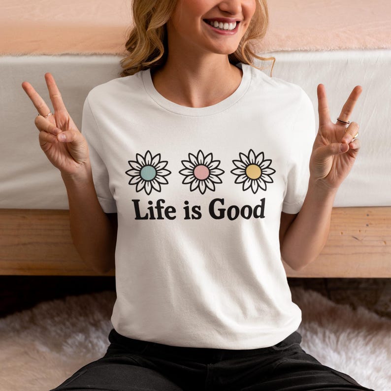 May include: White t-shirt with three daisy flower designs in pastel shades above the text "Life is Good". The flowers have blue, pink, and yellow centres. A casual, everyday wear item.
