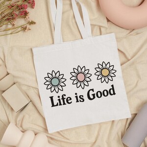 May include: White tote bag featuring three daisy flower designs in pastel colours and the text "Life is Good" in black. The bag has white handles and is on a beige fabric surface.