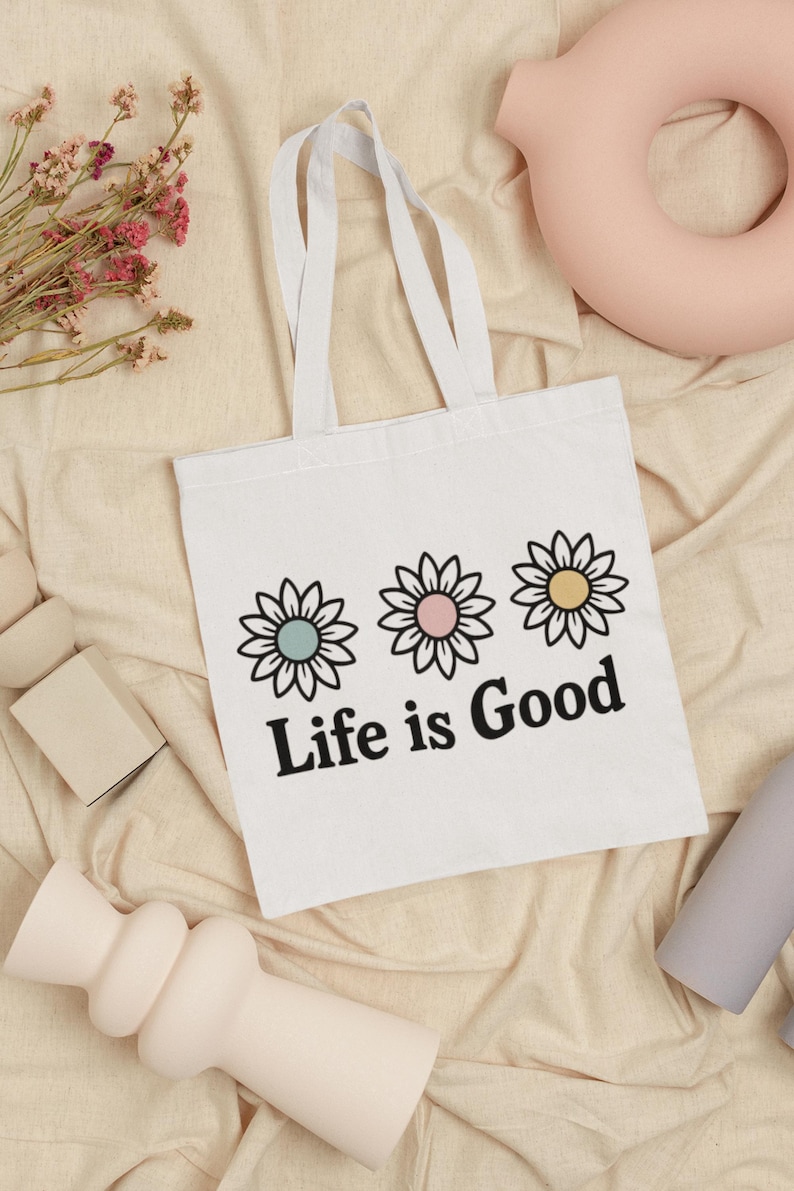 May include: White tote bag featuring three daisy flower designs in pastel colours and the text "Life is Good" in black. The bag has white handles and is on a beige fabric surface.
