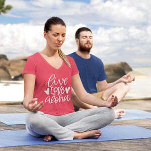 May include: Two people are sitting in a yoga pose on blue yoga mats. The woman in the foreground is wearing a red v-neck t-shirt with the text "live love aloha" in white. The man in the background is wearing a blue t-shirt.