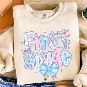 May include: Cream-colored sweatshirt with the words "First Grade" in a colorful, patterned design. The text features blue and pink gingham, floral, and bow accents, with a pencil and bow graphic at the bottom.