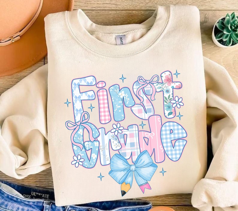 May include: Cream-colored sweatshirt with the words "First Grade" in a colorful, patterned design. The text features blue and pink gingham, floral, and bow accents, with a pencil and bow graphic at the bottom.