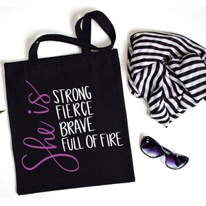 May include: Black tote bag with white text that reads "She is strong fierce brave full of fire". The text "She is" is in purple.