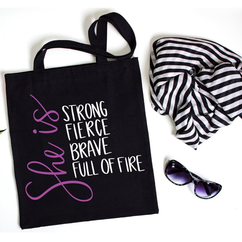 May include: Black tote bag with white text that reads "She is strong fierce brave full of fire". The text "She is" is in purple.
