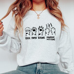 May include: A gray sweatshirt with a graphic of cat paws in the shape of rock, paper, scissors, and murder mittens. The text "Rock Paper Scissors Murder Mittens" is printed below the graphic.