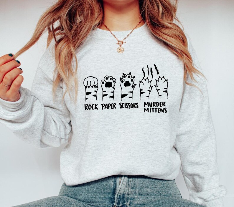 May include: A gray sweatshirt with a graphic of cat paws in the shape of rock, paper, scissors, and murder mittens. The text "Rock Paper Scissors Murder Mittens" is printed below the graphic.