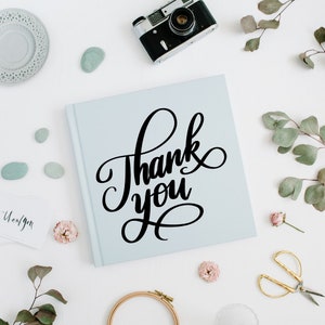May include: A light blue notebook with the words "Thank you" written in black calligraphy. The notebook is surrounded by decorative items, including a vintage camera, small stones, flowers, and a pair of gold scissors.